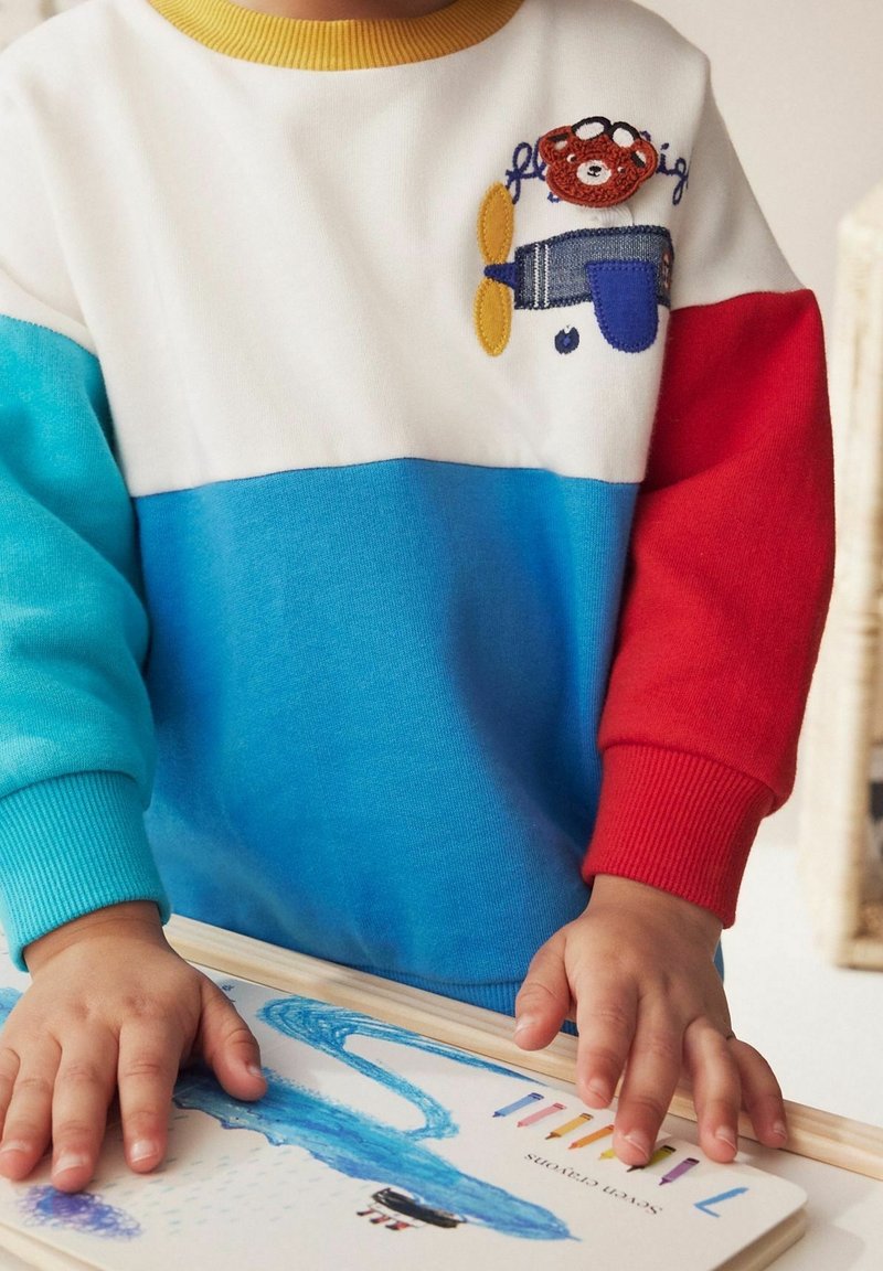 Enfant portant un pull à blocs de couleurs avec un écusson d'ours et d'avion, dessinant avec un crayon bleu sur une feuille avec des crayons de couleur à proximité.