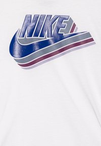 Camiseta blanca con un gran logo de Nike en azul y gris, con rayas rojas y moradas debajo del logo. Textura suave, diseño de manga corta.