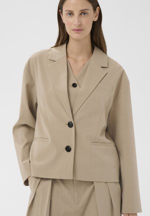 Naine, kes kannab beežikat ülisuurt bleiserit mustade nööpidega ja sobivaid voltidega pükse, seisab lihtsa hele tausta ees.