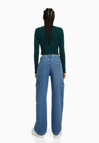 Zelený dlouhý rukáv s krátkým střihem a vroubkovaným lemem v kombinaci s širokými modrými džíny typu cargo s bočními kapsami a klasickým střihem.