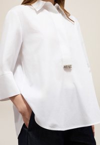 Blouse blanche surdimensionnée avec un col pointu, des manches trois-quart, des poches latérales et un accent décoratif argenté sur le devants.