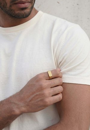 Mann trägt ein schlichtes weißes T-Shirt, hält und zieht leicht am Ärmel mit der linken Hand, trägt einen schlichten rechteckigen Goldring.