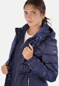 Giubbotto imbottito blu navy con cappuccio, texture trapuntata, chiusura con zip e polsini elasticizzati. Presenta una patch con logo sulla spalla e tasche laterali.