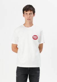 T-shirt bianco in cotone a maniche corte, collo rotondo e grafica circolare rossa con la scritta "981 REPLAY" sul lato sinistro del petto.