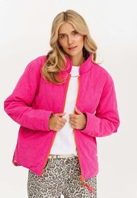 Różowa kurtka z korduroju z wysokim kołnierzem, jasnopomarańczowym zamkiem błyskawicznym i akcentami w panterkę na dolnej krawędzi. Noszona na białej bluzce.