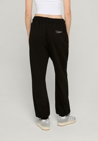 Svarta bomullssvettsbyxor med lös passform, elastiskt midjeband och vit logo broderad på baksidan. Har på sig grå sneakers.