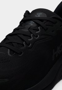 Κοντινή λήψη ενός μαύρου αθλητικού παπουτσιού με υφασμάτινο πλέξιμο, μαύρα κορδόνια και διακριτικές επωνυμίες στη γλώσσα και την πλευρά.