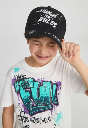 Cappello nero con il testo "CAMBIA LE REGOLE" in bianco, indossato da un ragazzo in una maglietta bianca con testo in stile graffiti colorato, con accenti blu e viola.