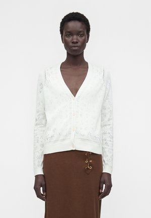 Donna che indossa un cardigan bianco con bottoni e motivi delicati, e una gonna marrone con dettagli ad anello dorati, in piedi davanti a uno sfondo semplice.