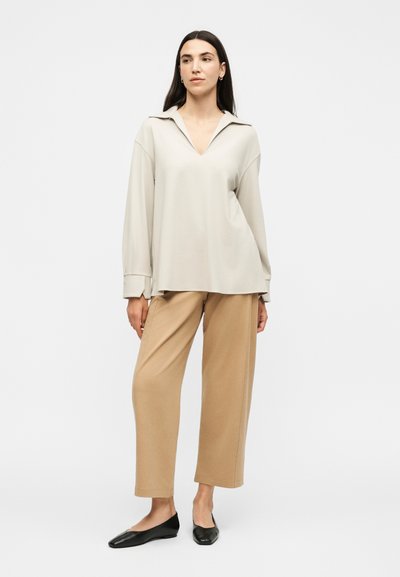 Γυναίκα με μακριά σκουρόχρωμα μαλλιά, που φοράει ένα άνετο μπεζ μακρυμάνικο μπλουζάκι, καφέ παντελόνι με φαρδιά μπατζάκια και μαύρα παπούτσια χωρίς τακούνια, στέκεται μπροστά σε ένα απλό φόντο.