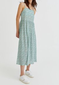 Vestido sin mangas de estampado floral en verde, con un cuerpo ajustado y cintura fruncida, que cae a media pantorrilla. Combinado con zapatillas blancas.