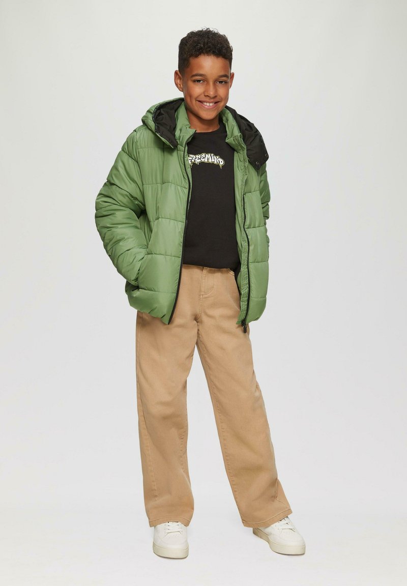 Grüne Puffjacke mit schwarzer Kapuze, schwarzes T-Shirt mit Grafik, beige weite Hose und weiße Turnschuhe, präsentiert vor einem schlichten Hintergrund.