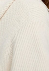 Strickpullover in Cremefarbe mit Rippstruktur, hohe Kragenform und diagonalen Nahtdetails an der Schulter.