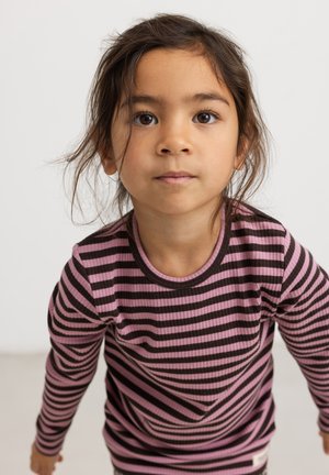 Ung pige med brune øjne og løst mørkt hår iført en langærmet, lyserød og sort stribet skjorte, der kigger lige frem.