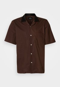 Chemise marron à manches courtes avec des boutons, un col noir et une poche sur la poitrine, présentée sur un fond blanc uni.