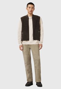 Brown fleece vest with side zippers over a light beige sweatshirt. Beige pants with patchwork detail, paired with textured black shoes.