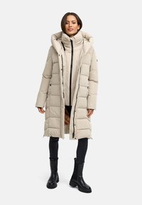 Beige langer Puffer-Mantel mit hohem Kragen und Reißverschluss vorne. Verfügt über ein gestepptes Design und schwarze Akzente. Kombiniert mit schwarzen Jeans und Stiefeln.