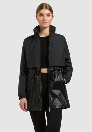 Mladá žena v čiernej bunde s vysokým golierom a zipsom, s vreckami so zúženým pásom, stojaca na pozadí s jednofarebnou svetlošedou stenou.