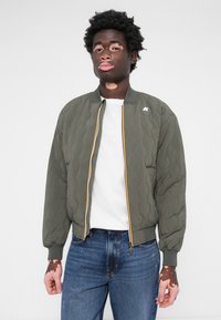 Töltött bomber dzseki olíva zöld színben, sárga és fehér csíkokkal, bordás mandzsettákkal és cipzáras záródással. Fehér póló felett viselve.