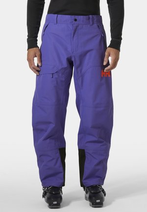 Hombre vistiendo pantalones de esquí morados con puños reforzados negros y botas de esquí negras, de pie contra un fondo blanco.