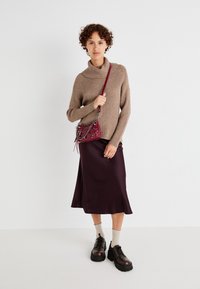 Maglione marrone a coste con collo a scialle, gonna midi in satin bordeaux e scarpe con tacco piattaforma nere con lacci. Porta una piccola borsa a spalla bordeaux.