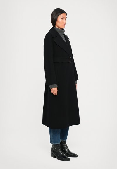 Femme portant un long manteau noir ceinturé sur un col roulé gris, un jean bleu et des bottines noires à talons avec des œillets argentés, se tenant de profil.