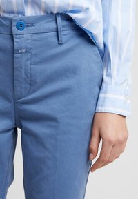 Gros plan sur une personne portant un pantalon bleu clair avec un bouton bleu et une chemise à rayures bleu clair à manches longues avec un bouton aux poignets.