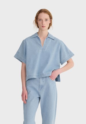 Młoda kobieta o jasnej karnacji i rudych włosach, ubrana w jasnoniebieską, oversize'ową, krótką koszulę z denimu oraz dopasowane, wysokie dżinsy, z ręką w kieszeni.