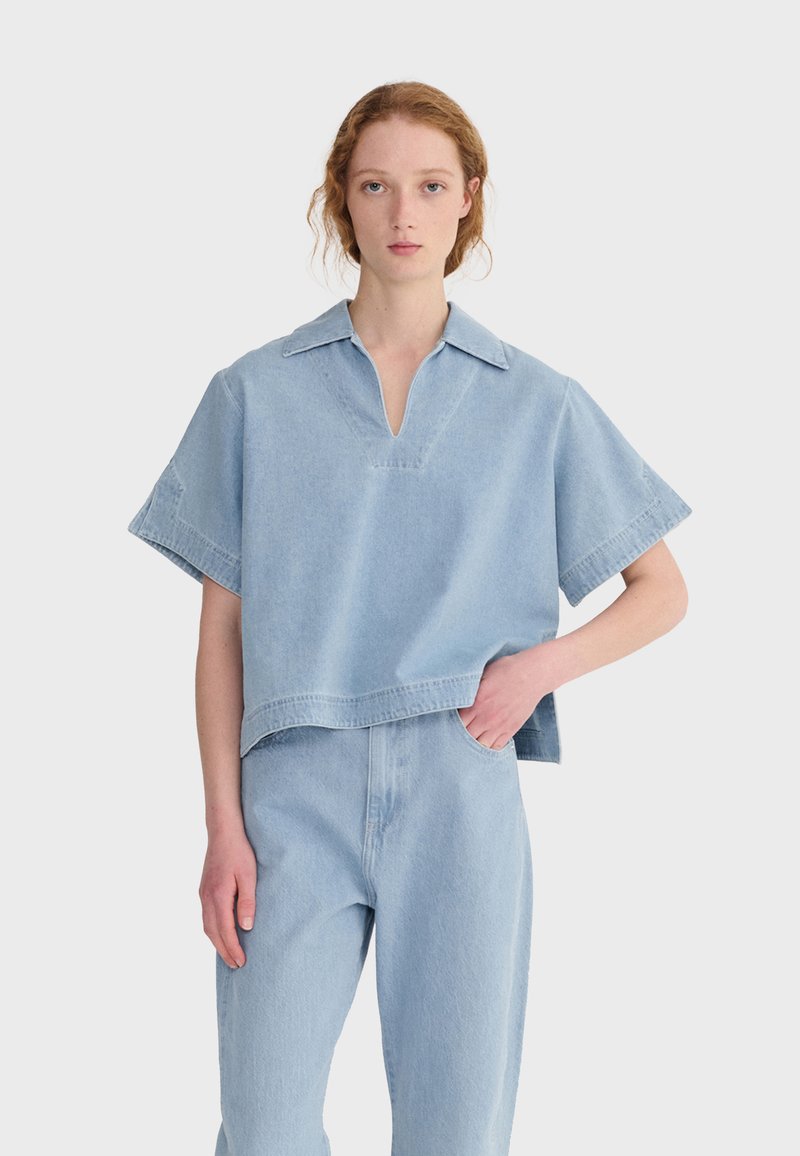 Jeune femme à la peau claire et aux cheveux roux portant une chemise en jean bleu clair à manches courtes oversize et un jean taille haute assorti, main dans la poche.