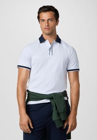 Biela polokošeľa s námorníckym golierom a akcentmi na rukávoch. Má zips v oblasti krku a tmavozelený sveter zaviazaný v páse.