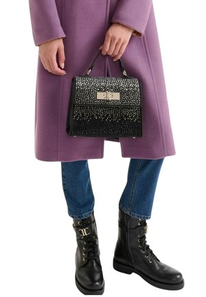 Borsa nera con borchie argentate, forma rettangolare e chiusura metallica, tenuta da una persona con cappotto lilla e jeans blu, abbinata a stivali neri.