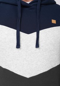 Hanorac cu glugă în nuanțe de bleumarin, alb și gri. Prezintă un model în formă de săgeată, șnururi și un mic logo pătrat de culoare bej pe piept.