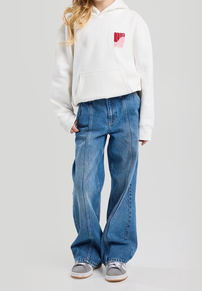 Hoodie en coton blanc avec un logo rouge, associé à un jean en denim bleu à jambes larges. Des baskets grises complètent le look, arborant un tissu texturé.