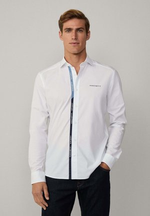 Camisa blanca de botones con un panel lateral azul en contraste que presenta el texto "HACKETT SPORT". Mangas largas y un diseño de cuello clásico.