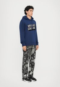 Felpa blu navy con logo "Versace Jeans Couture"; pantaloni neri a motivo con design floreali e ornamentali; sneakers nere.