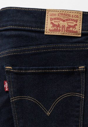 Jean en denim bleu foncé avec un patch en cuir au niveau de la taille indiquant la marque. Comprend des coutures jaunes et une étiquette rouge sur la poche arrière.