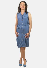 Vestido sin mangas azul con botones en el frente, dos bolsillos en el pecho y un lazo en la cintura. Tejido suave con un corte holgado; combinado con tacones altos azules.