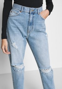 Ljusblå högmidjade slitna jeans med hål vid knäna och låret, kombinerat med en svart långärmad topp, med en hand i fickan.