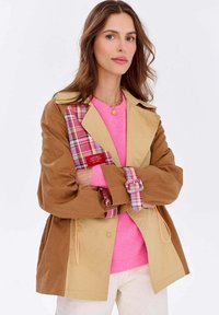 Hnedo-zelená dlhá bunda s kockovaným akcentom, v kombinácii s jasne ružovým sveterom. Obsahuje gombíkové manžety a predné vrecká.