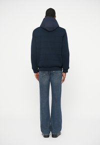 Ναυτικό ριμπ πουλόβερ με κουκούλα, που διαθέτει υφαντές πάνελ. Συνδυάζεται με ανοιχτόχρωμο μπλε φαρδύ τζιν και μαύρα παπούτσια, με θέα από πίσω.