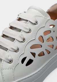 Λευκό δερμάτινο αθλητικό παπούτσι με φαρδιές υφασμάτινες κορδόνια και λουλουδάτα σχέδια κοπής στο πλάι, που αποκαλύπτουν ταμπά εσωτερική επένδυση.