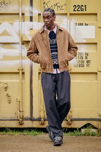 Barna szarvasbőr bombázó dzseki tengerészkék, fehér nyomattal díszített pulóver felett, kockás ing és tűsávos nadrág. Fekete fűzős cipő egészíti ki az öltözéket.