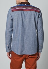 Camisa azul de chambray con un tapete estampado en tonos rosas y azules, cuello marrón y mangas largas, con puños abotonados y detalle de costura en la espalda.