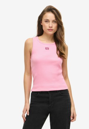 Naine, kellel on pikad lainelised juuksed, kannab varrukateta roosat topi väikese punase logoga ja musti kõrge vöökohaga teksaseid, seisab valge tausta ees.