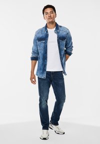 Ljusblå denimjacka med två bröstfickor, bärs över en vit t-shirt, matchad med mörkblå jeans och vita sneakers.