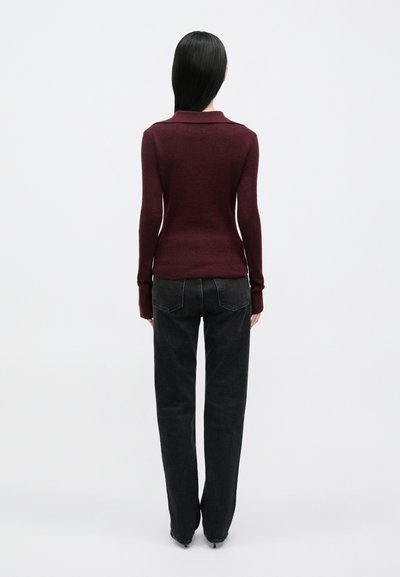 Γυναίκα με μακριά μαύρα μαλλιά με την πλάτη στραμμένη, φορώντας μπορντό μπλούζα με μακριά μανίκια και μαύρο τζιν με ίσια γραμμή και μαύρες γόβες.