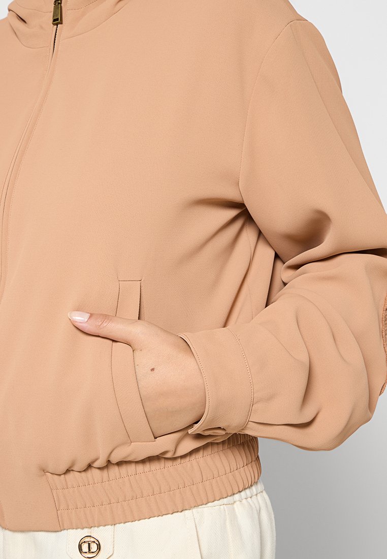 Chaqueta cremosa con cremallera, cintura elástica, mangas acampanadas y mano en el bolsillo delantero izquierdo, sobre pantalones blancos con detalle de botones.