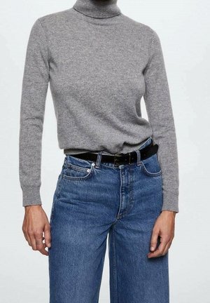 Personne portant un pull à col roulé gris rentré dans un jean bleu taille haute avec une ceinture noire sur un fond uni.