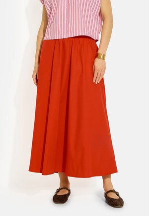 Mujer que lleva una falda roja hasta los tobillos, una camisa rosa a rayas con botones, zapatos planos con estampado de leopardo y una pulsera de puño dorada en la muñeca izquierda.