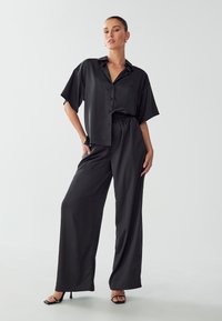 Camicia nera in satin a maniche corte con bottoni e pantaloni neri a vestibilità ampia con cintura elastica, abbinati a sandali neri con tacco alto.